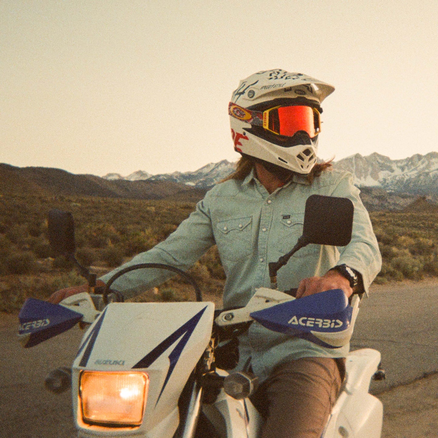 A person in a rugged denim shirt, white helmet, and goggles sits on a Suzuki motorcycle, looking to the side. Mountains and open landscape stretch behind under a clear sky, highlighting this adventurous and versatile style.