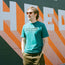 A man with light brown hair, sunglasses, and a teal Howler Brothers T-shirt stands against a colorful mural featuring bold geometric shapes and large orange and white letters, echoing the vibe of experimental artwork. His hands are in his pockets.