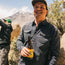 A man in a rugged cotton work shirt and cap smiles while holding a beer bottle outdoors, with mountains and greenery in the background. Another person, partially visible, stands to the left adjusting a camera or gear.