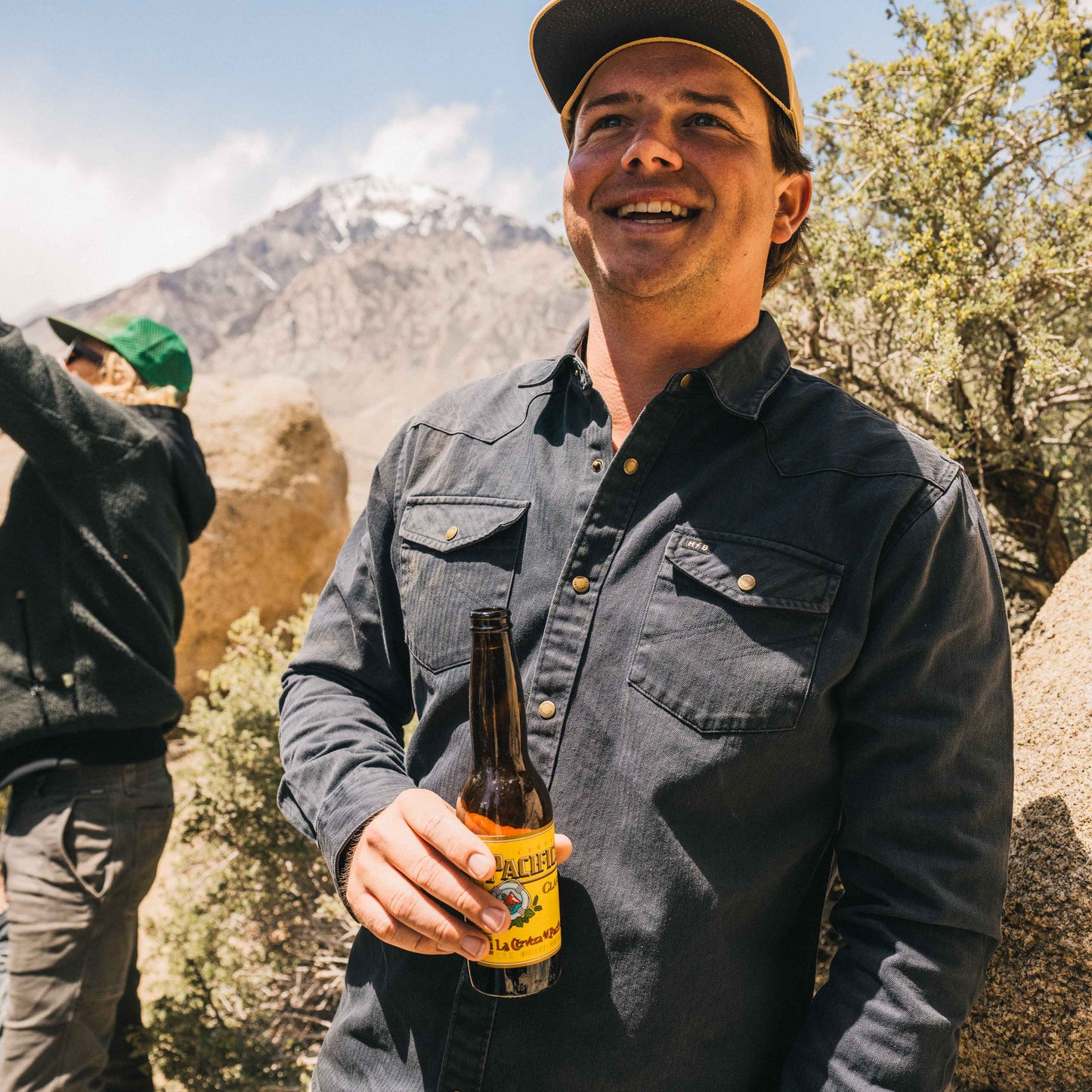A man in a rugged cotton work shirt and cap smiles while holding a beer bottle outdoors, with mountains and greenery in the background. Another person, partially visible, stands to the left adjusting a camera or gear.