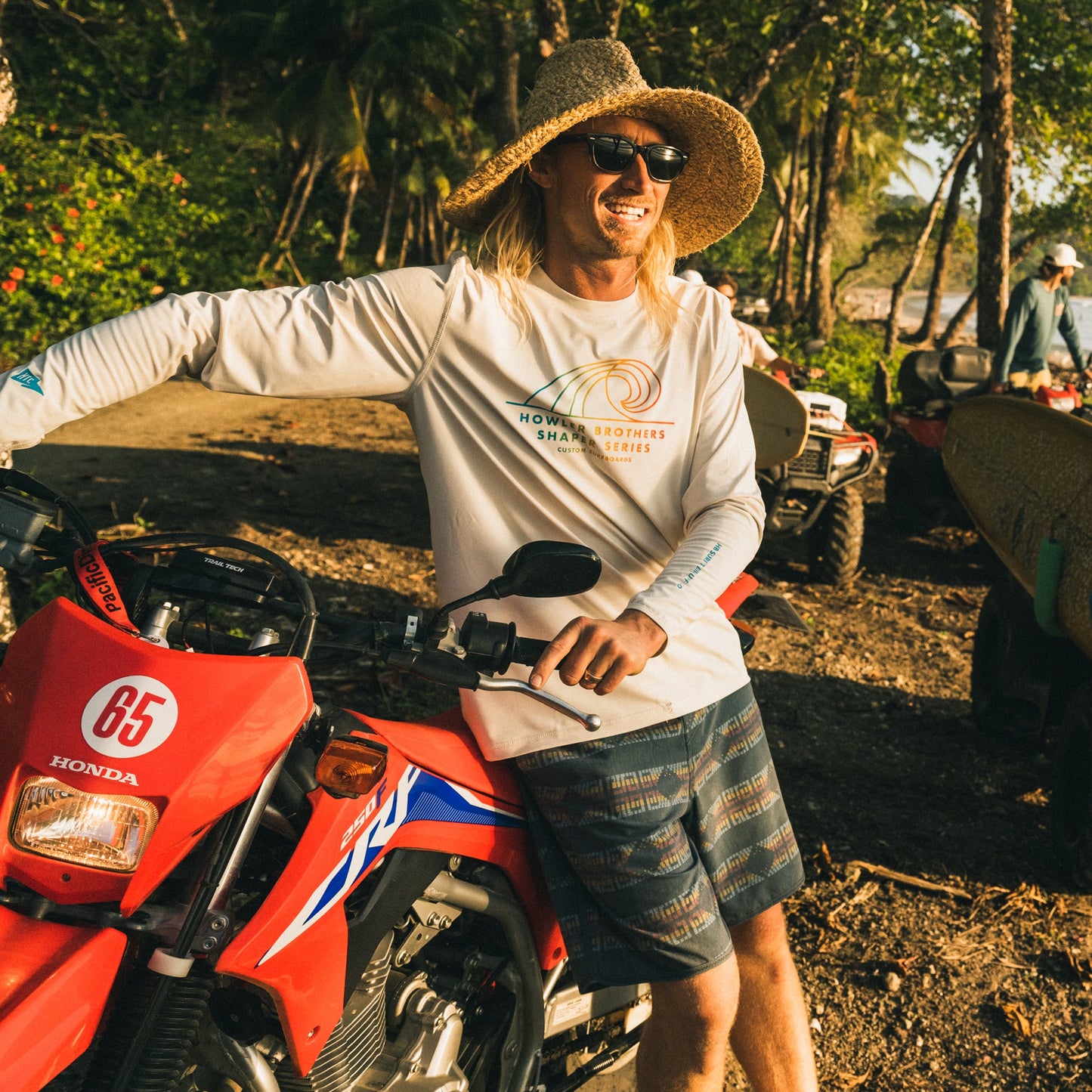 A man wearing a straw hat, sunglasses, and a light long-sleeve shirt stands outdoors, smiling and leaning on a red Honda ATV in a sunny, tropical setting with palm trees—perfect for showing off his Bruja Boardshorts.