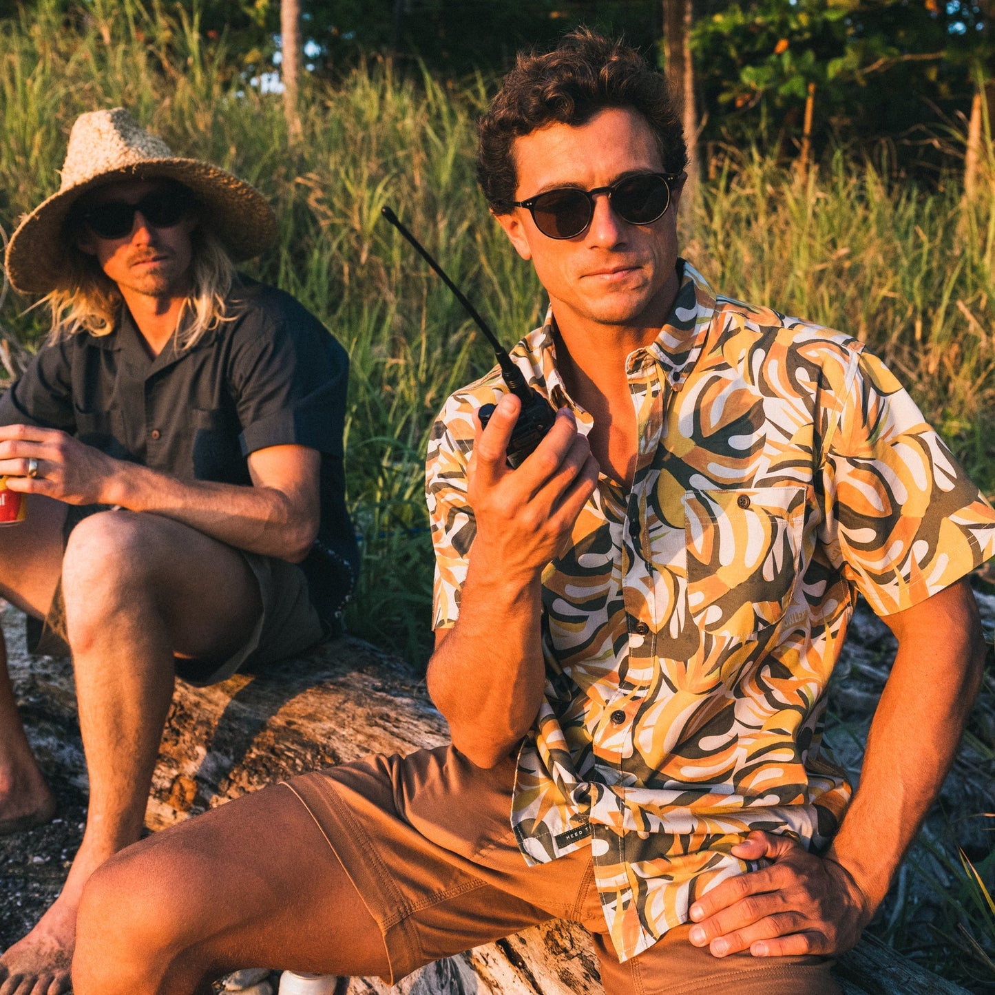 Two men sit outdoors on a log; one sports a patterned Mansfield Shirt and sunglasses, holding a walkie-talkie, while the other wears a straw hat and sunglasses with a drink in hand. Grass and trees form the background for this relaxed scene.