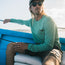 A man wearing sunglasses, a cap, and a green Howler Brothers UPF 50+ shirt sits on a blue boat holding fishing equipment, looking off to the side in bright sunlight—ready for any outdoor activities.