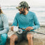 A man in a blue long-sleeve shirt and soft crown camouflage cap smiles while sitting on a log at the beach, holding a fishing rod and a white mug. Waves and sand are visible in the background.