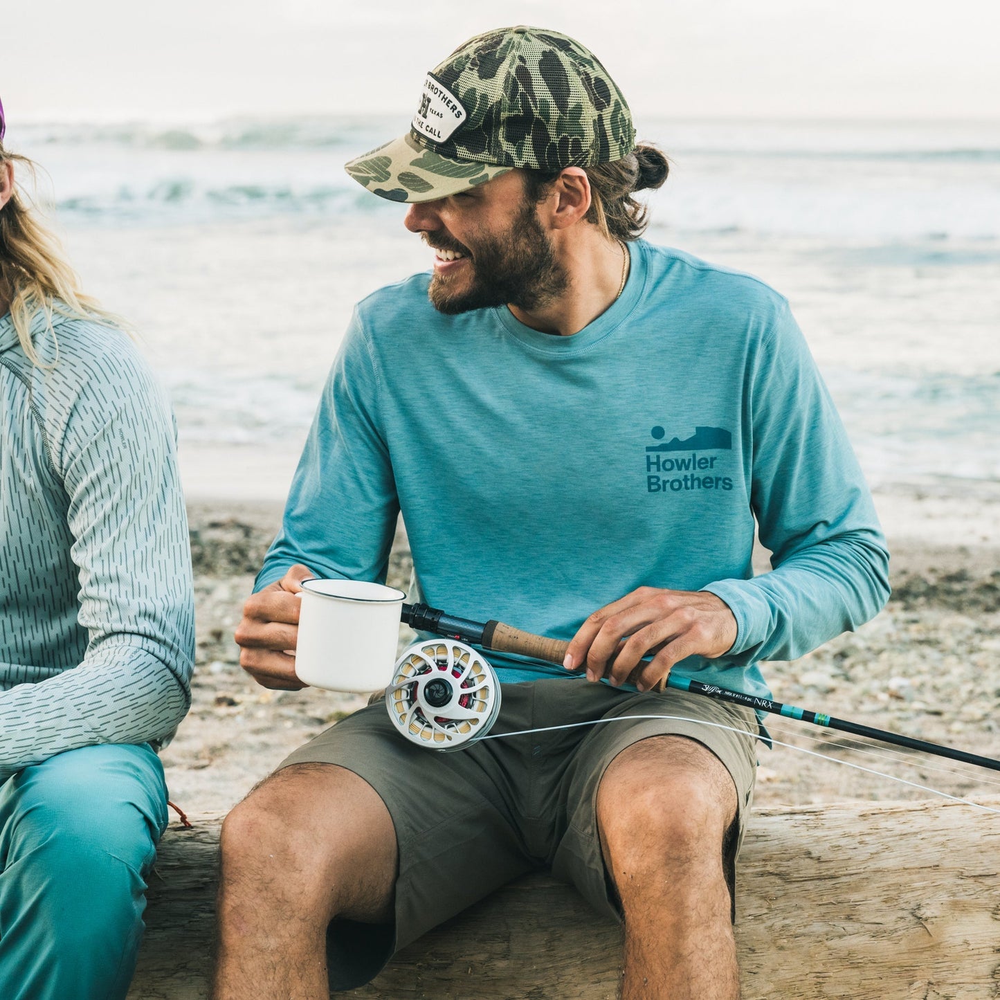 A man in a blue long-sleeve shirt and soft crown camouflage cap smiles while sitting on a log at the beach, holding a fishing rod and a white mug. Waves and sand are visible in the background.