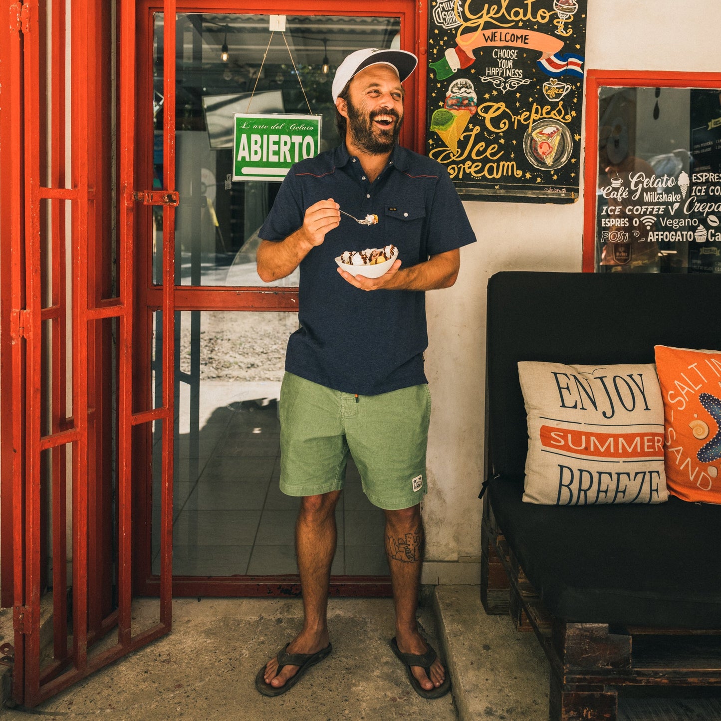 A man in Pressure Drop Cord Shorts, a polo shirt, and a cap stands by a red door, smiling and eating ice cream. Behind him are colorful signs and pillows on a bench that say “Enjoy Summer Breeze.”.