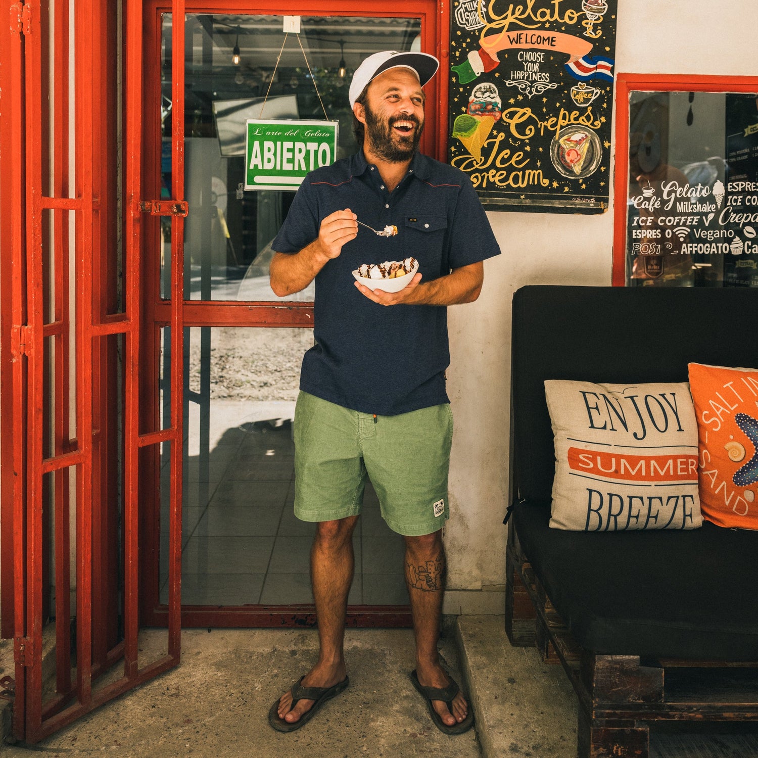 A man in Pressure Drop Cord Shorts, a polo shirt, and a cap stands by a red door, smiling and eating ice cream. Behind him are colorful signs and pillows on a bench that say “Enjoy Summer Breeze.”.