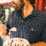 A bearded man in a 100% cotton navy blue Ranchero Polo with western yokes holds a bowl of ice cream topped with chocolate syrup, about to enjoy a spoonful. His face is partially out of frame.