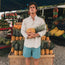 A man in a light shirt and quick drying nylon shorts stands outside at a fruit stand, holding a pineapple. He is surrounded by pineapples and other produce, with colorful fruit displays in the background.