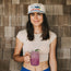 A woman in a light crop top and an unstructured snapback with a soft crown holds a mason jar of purple lemonade with a lime slice, standing in front of wooden shingles, smiling at the camera.