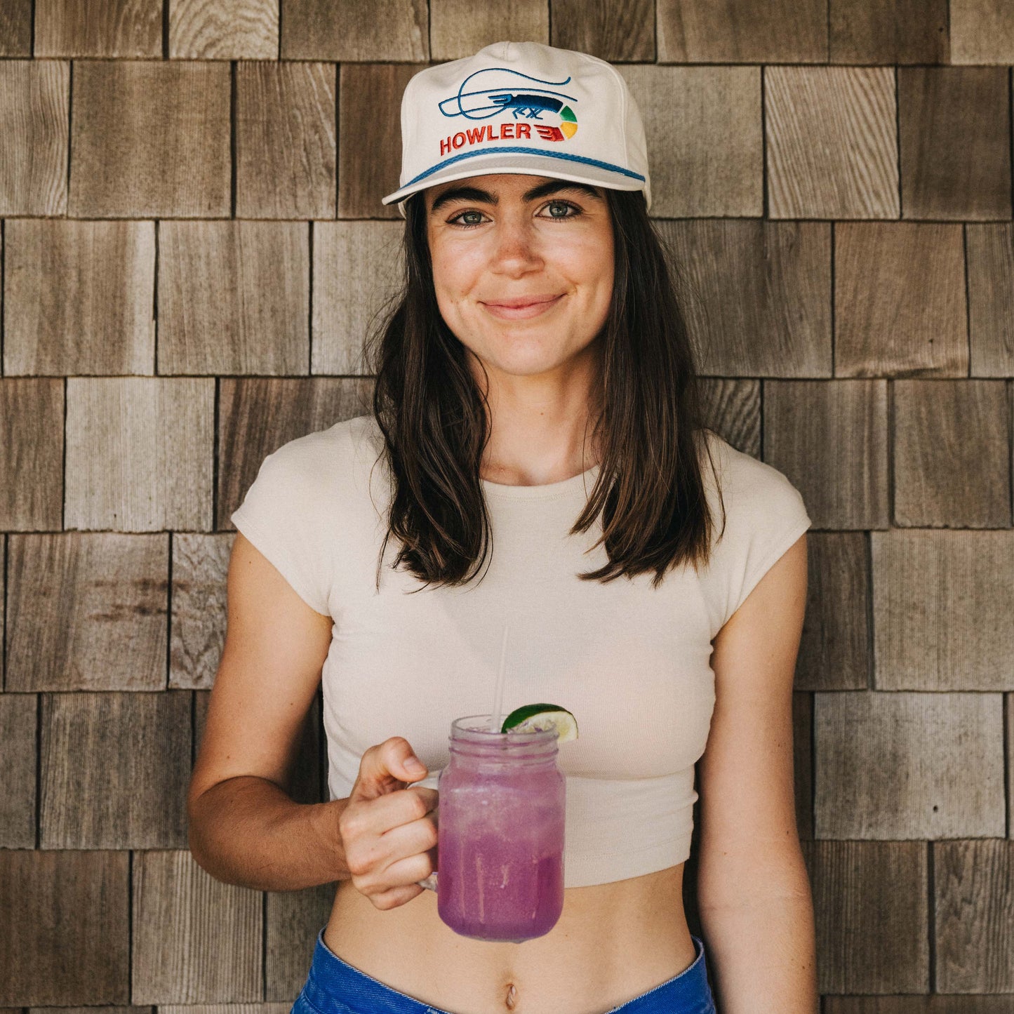 A woman in a light crop top and an unstructured snapback with a soft crown holds a mason jar of purple lemonade with a lime slice, standing in front of wooden shingles, smiling at the camera.