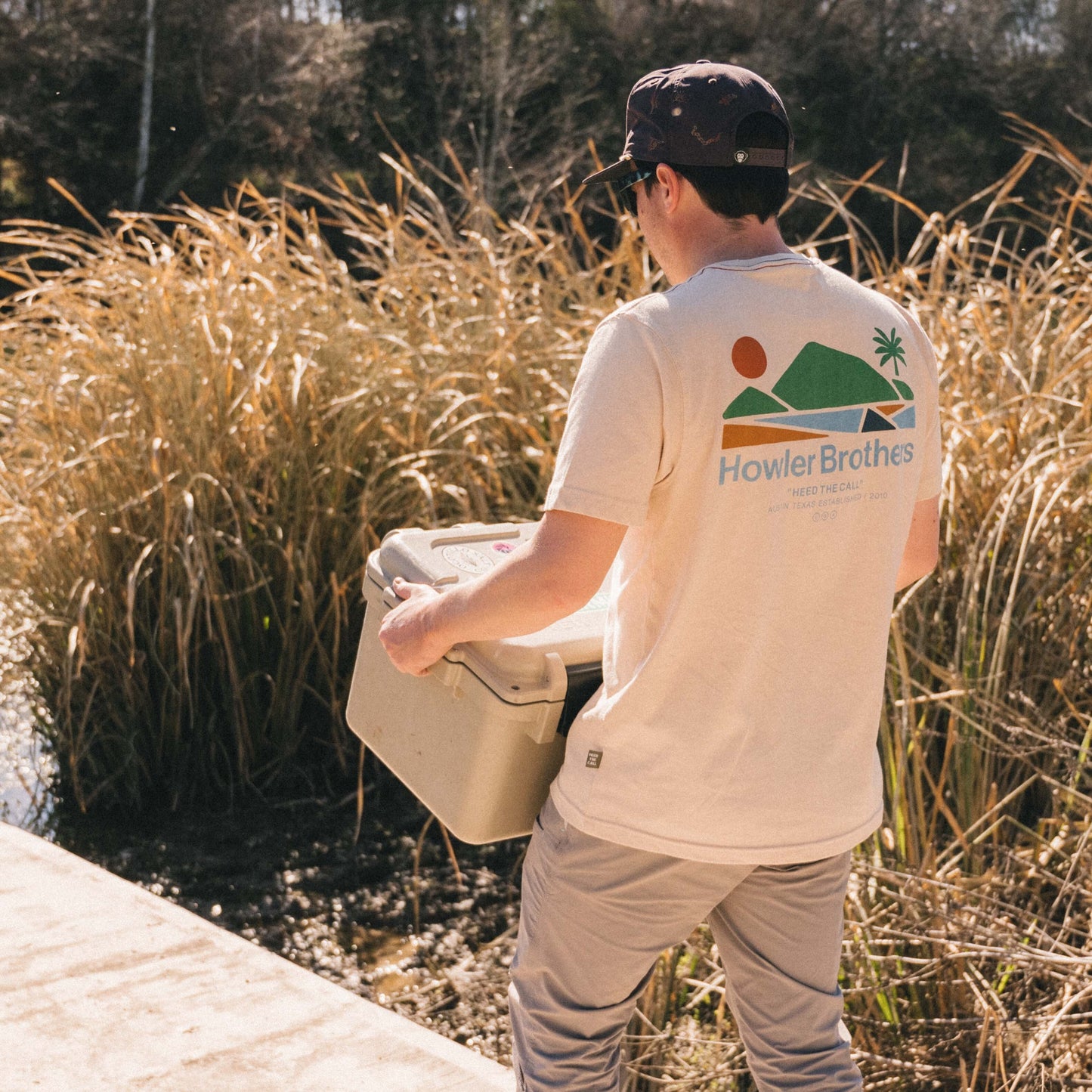 A man in a Howler blended t-shirt and cap carries a cooler near tall grass by the water on a sunny day, enjoying maximum comfort from the soft cotton-poly blend.