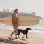 A man with long blond hair, wearing quick drying boardshorts, carries a surfboard while walking on a sandy beach next to a brown dog, with ocean waves and cliffs in the background.
