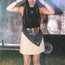 A woman in a black sleeveless top and beige skirt stands outdoors, smiling, with a Howler Bandanas scarf reading ACL Music Festival tied around her waist. She has long hair, bracelets, and a heart-shaped necklace.