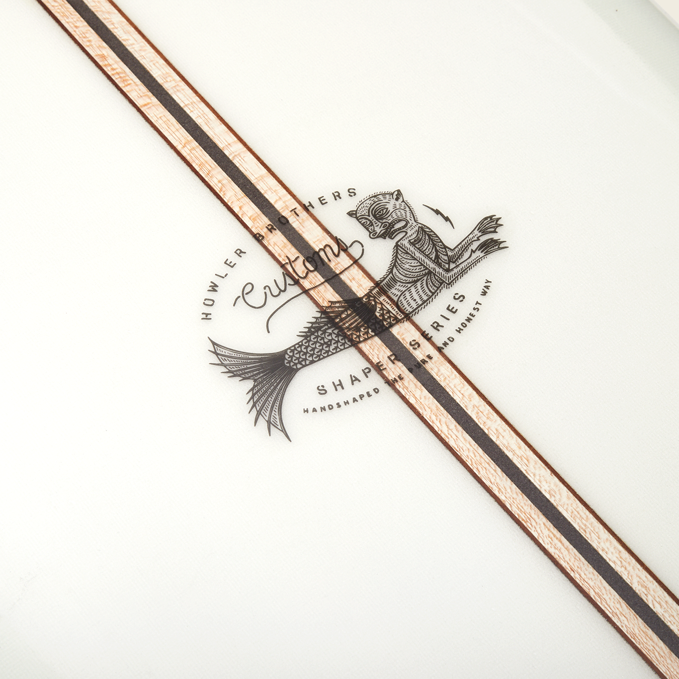 Close-up of a traditional surfboard featuring a logo with a fish and the words “Howler Brothers Custom Shapes Series,” plus two parallel wood inlays running diagonally across the white surface, reminiscent of classic noseriding boards.