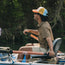 A person wearing a vintage-inspired hat and sunglasses sits in a boat, holding oars, rowing on a calm body of water surrounded by green foliage.