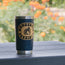 A navy blue YETI tumbler with a Monosonic Sessions and Howler Brothers sticker sits on a gray wooden table, with blurred orange flowers and greenery in the background.