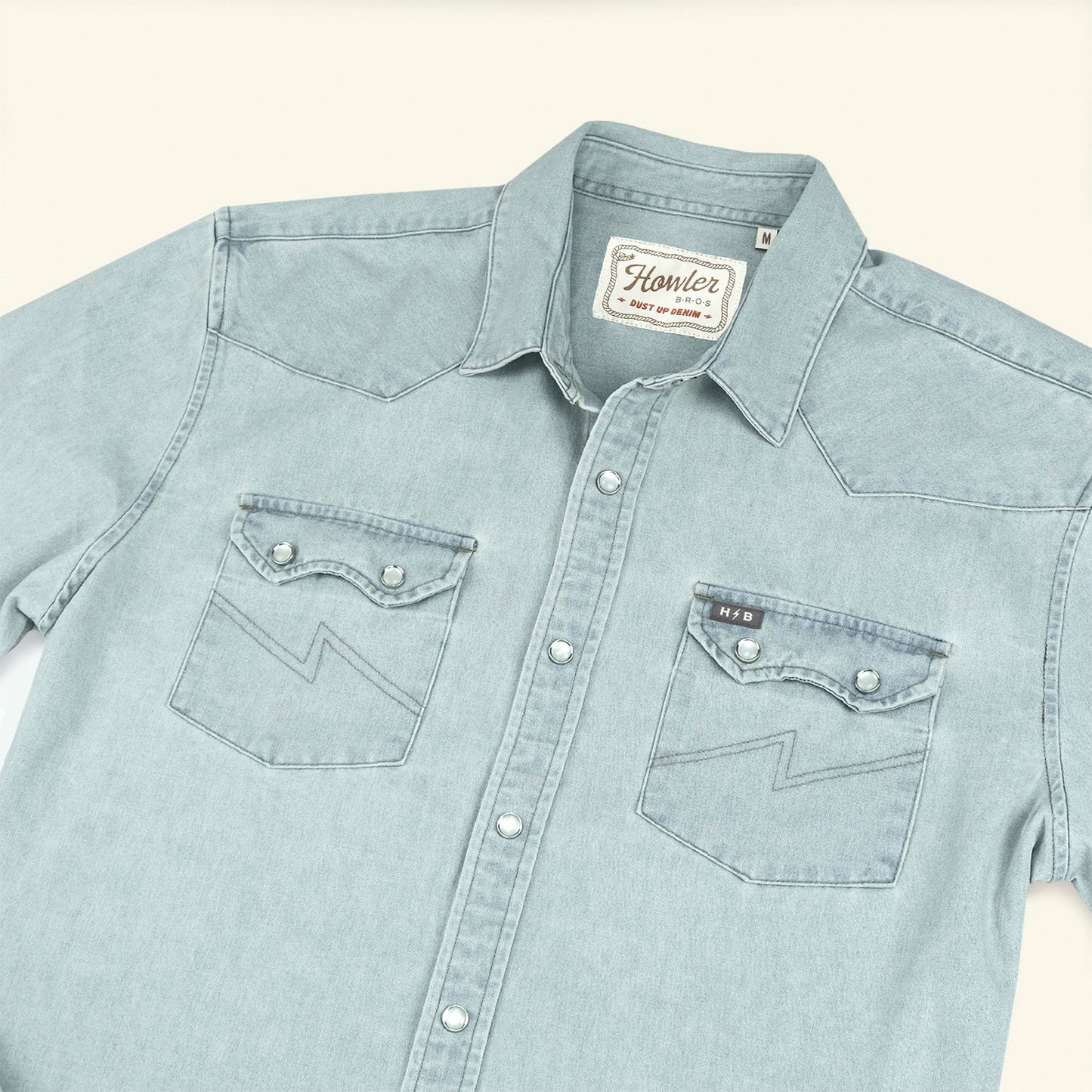 A light blue Denim Snapshirt with rugged denim construction, two chest pockets featuring pointed flaps and zigzag stitching, silver snap buttons, and a Howler brand label on the inside collar—perfect for versatile style.