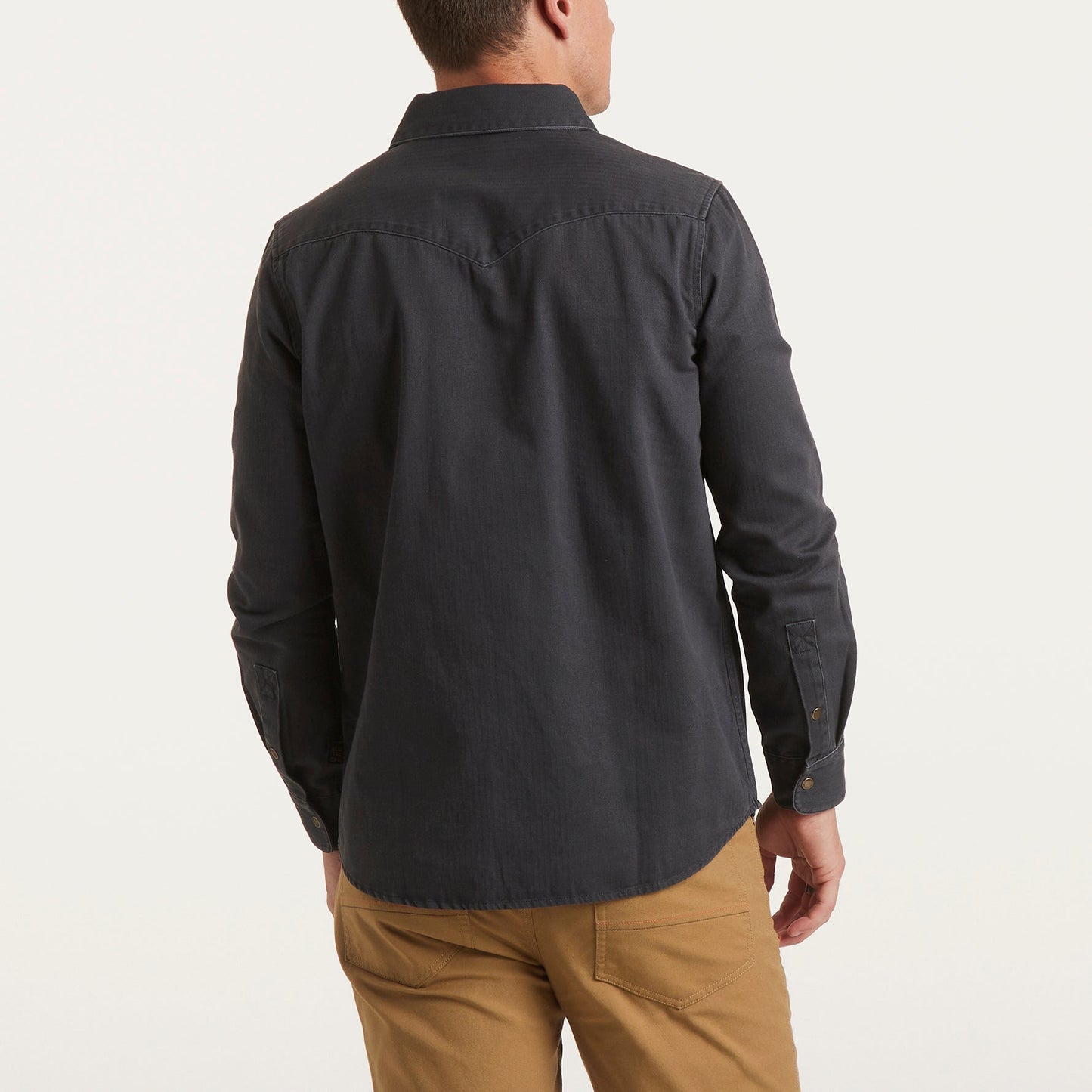 A man is standing with his back to the camera, wearing a dark Sawhorse Work Shirt and light brown pants against a plain, light background.