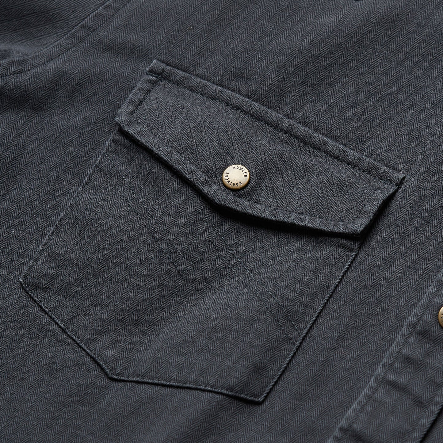 Close-up of a dark gray herringbone Sawhorse Work Shirt, featuring a buttoned flap chest pocket with visible stitching and a silver metal button.