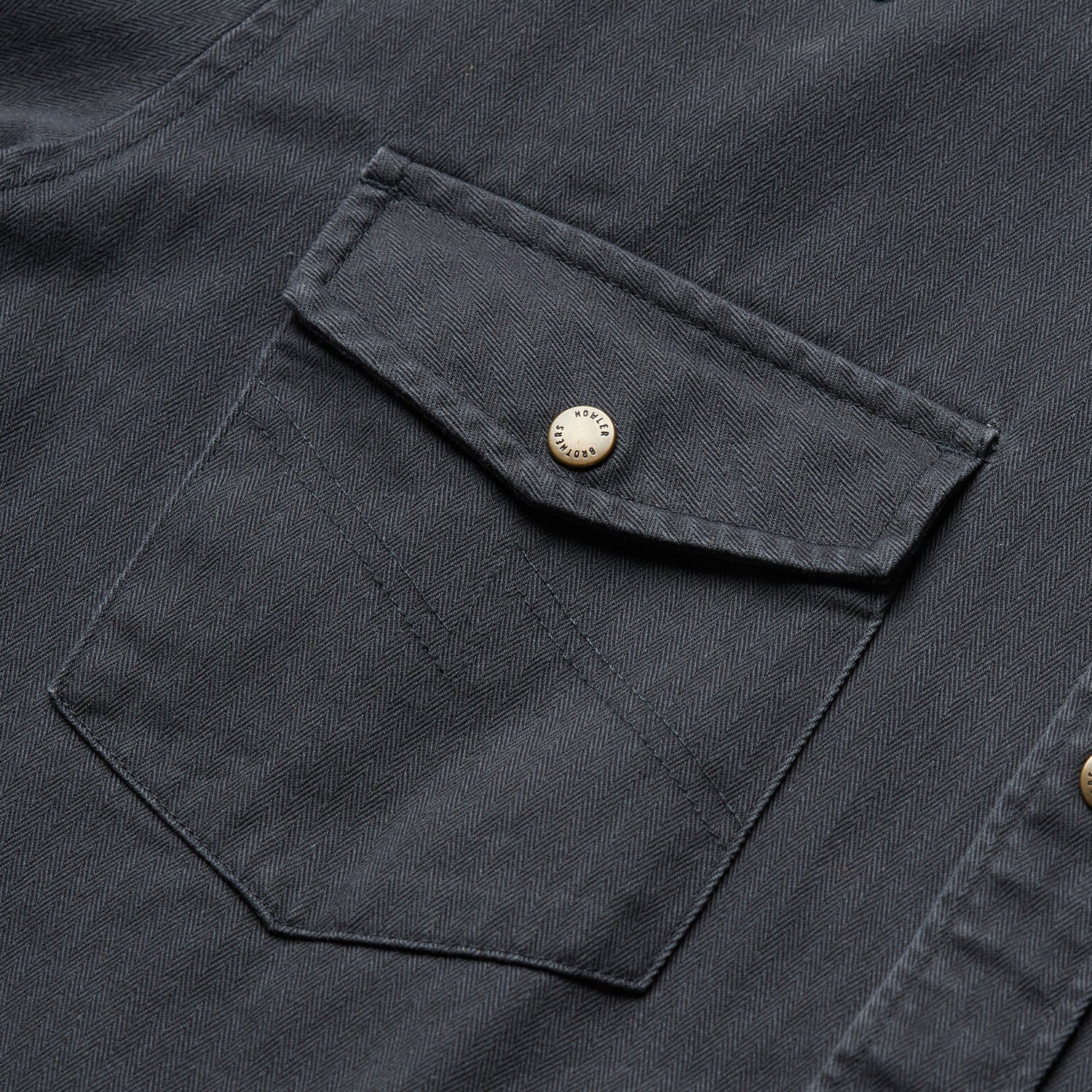 Close-up of a dark gray herringbone Sawhorse Work Shirt, featuring a buttoned flap chest pocket with visible stitching and a silver metal button.