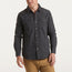 A man wearing a rugged cotton Sawhorse Work Shirt with two chest pockets and tan pants stands against a plain, light background. The shirt features metallic buttons and a casual fit.