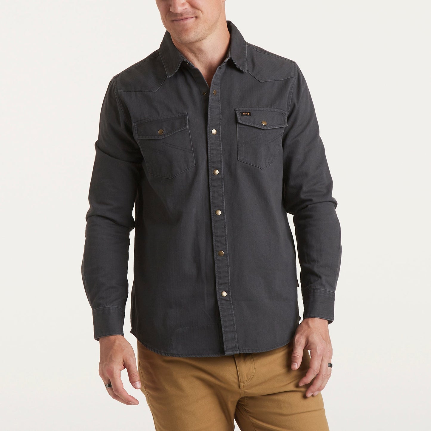 A man wearing a rugged cotton Sawhorse Work Shirt with two chest pockets and tan pants stands against a plain, light background. The shirt features metallic buttons and a casual fit.