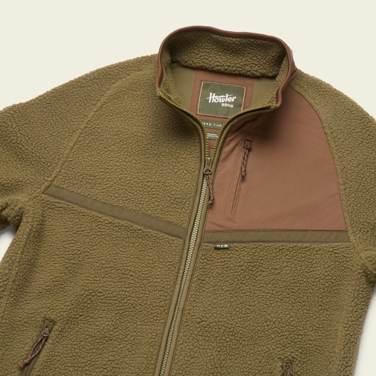 A brown polyester fleece jacket with a front zipper, high collar, and a chest pocket featuring a contrasting panel. The soft and fluffy texture adds comfort, while the label inside reads Howler Bros Howler Crozet.