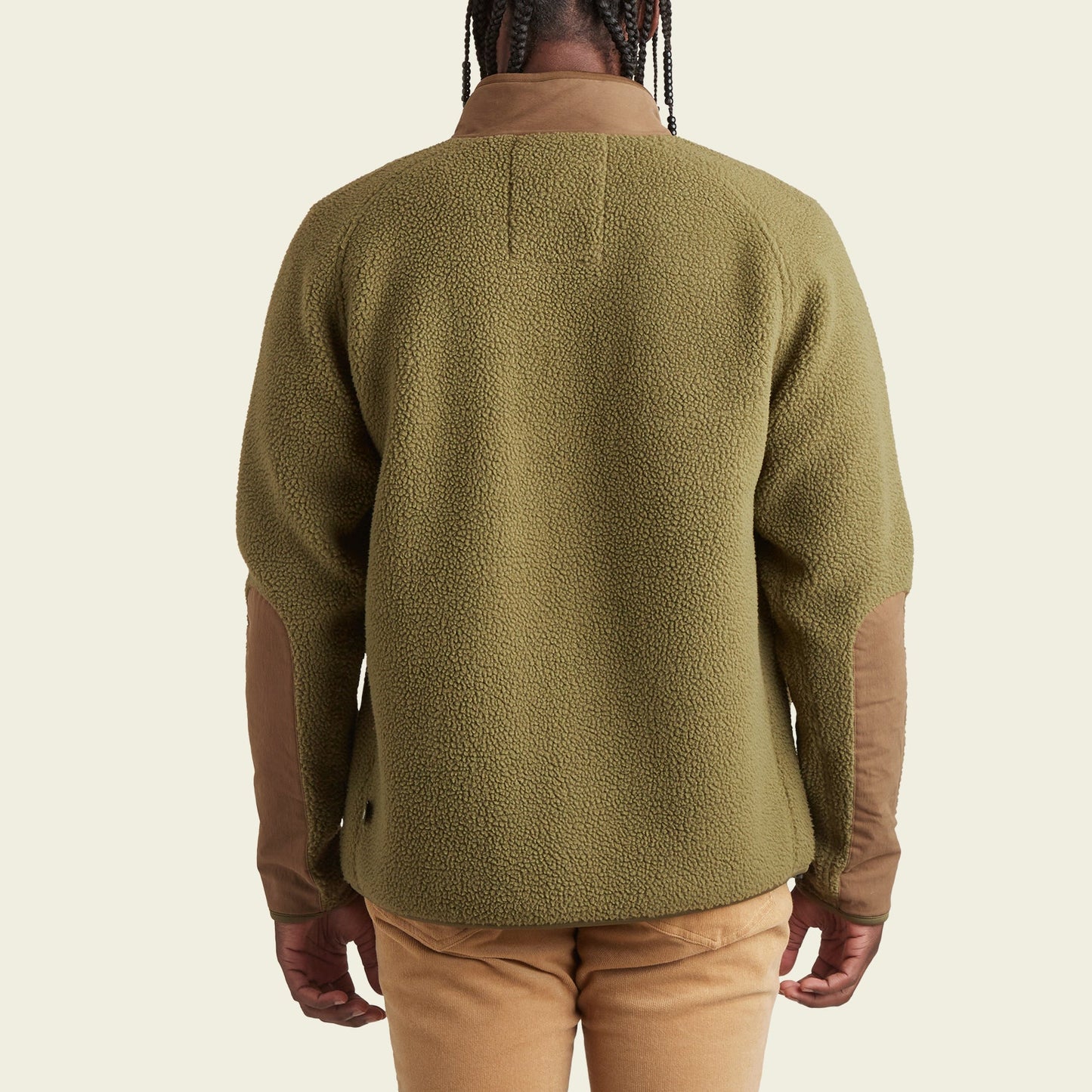 A person wearing a green Howler Crozet fleece jacket with brown elbow patches and tan pants is shown from the back against a plain light background.