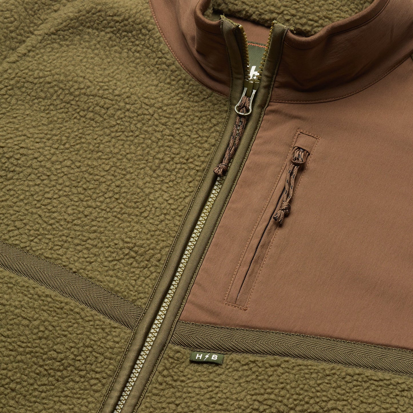 Close-up of an olive green and brown polyester fleece jacket with a zipper, a chest pocket featuring a zipper pull, and a small H/B tag near the main zipper. The soft, textured fabric makes it perfect for fall outerwear.