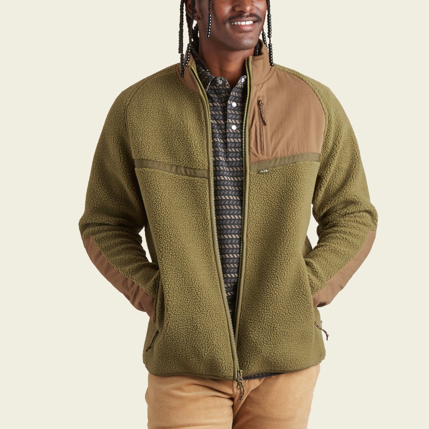 A person wearing an olive green fleece jacket with tan fabric accents on the shoulders and pockets, a patterned shirt underneath, and tan pants stands with hands in jacket pockets, smiling—ready for stylish fall outerwear.