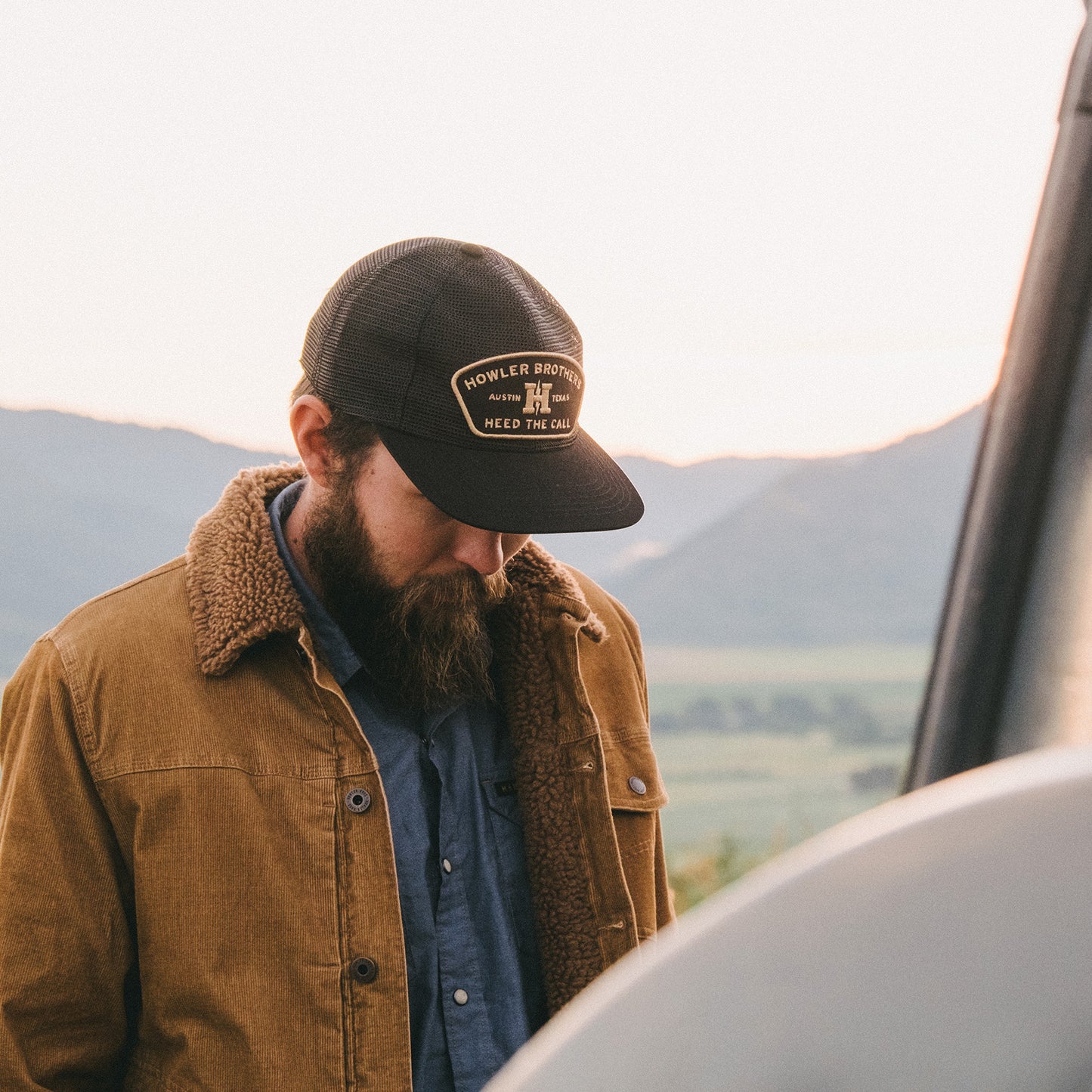 A bearded man in a black cap and corduroy jacket with sherpa lining looks down, standing outdoors with mountains and a soft, pastel-colored sky in the background.
