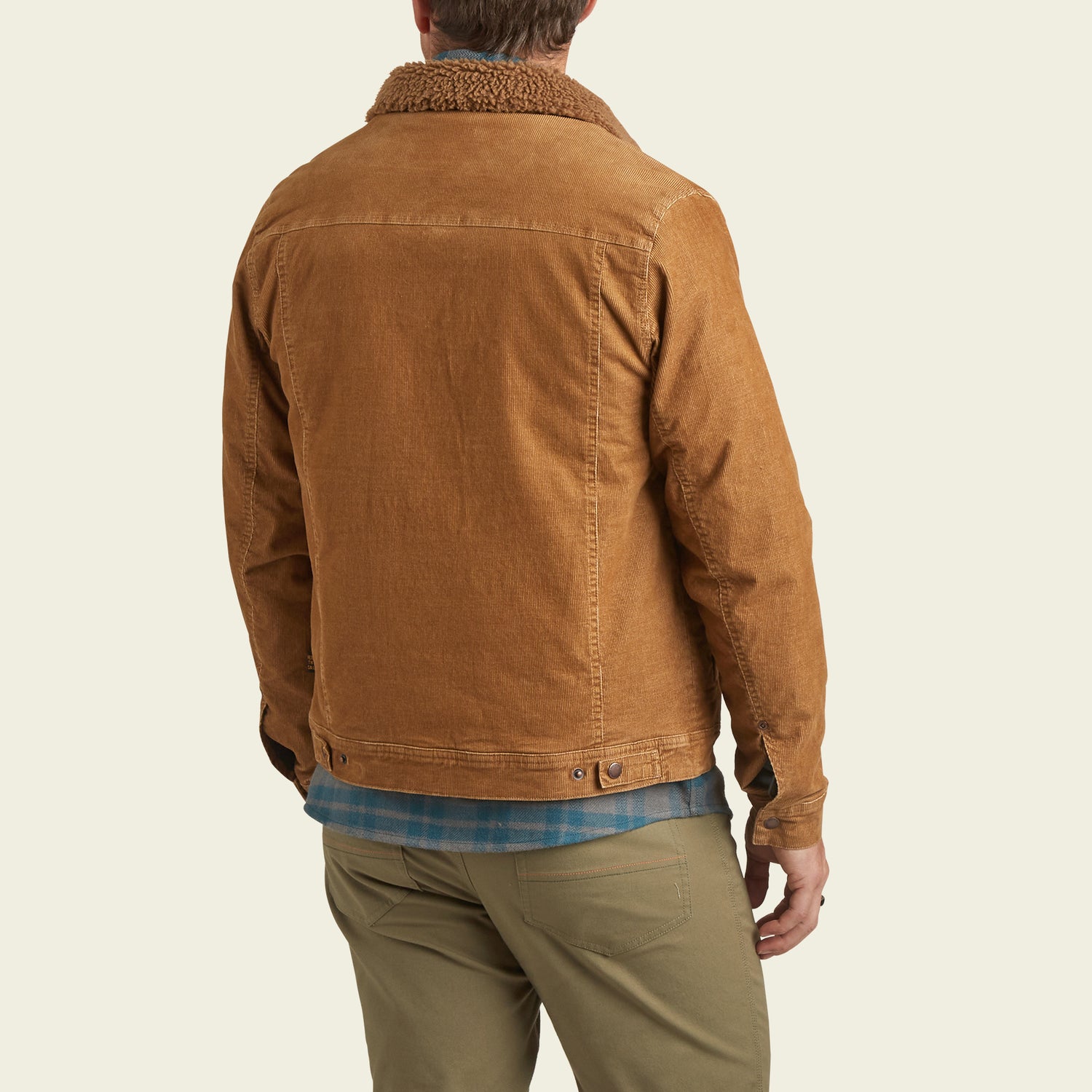 A person wearing the Fuzzy Depot Jacket—a light brown corduroy jacket with a sherpa lining—khaki pants, and a blue plaid shirt stands facing away from the camera against a light background.