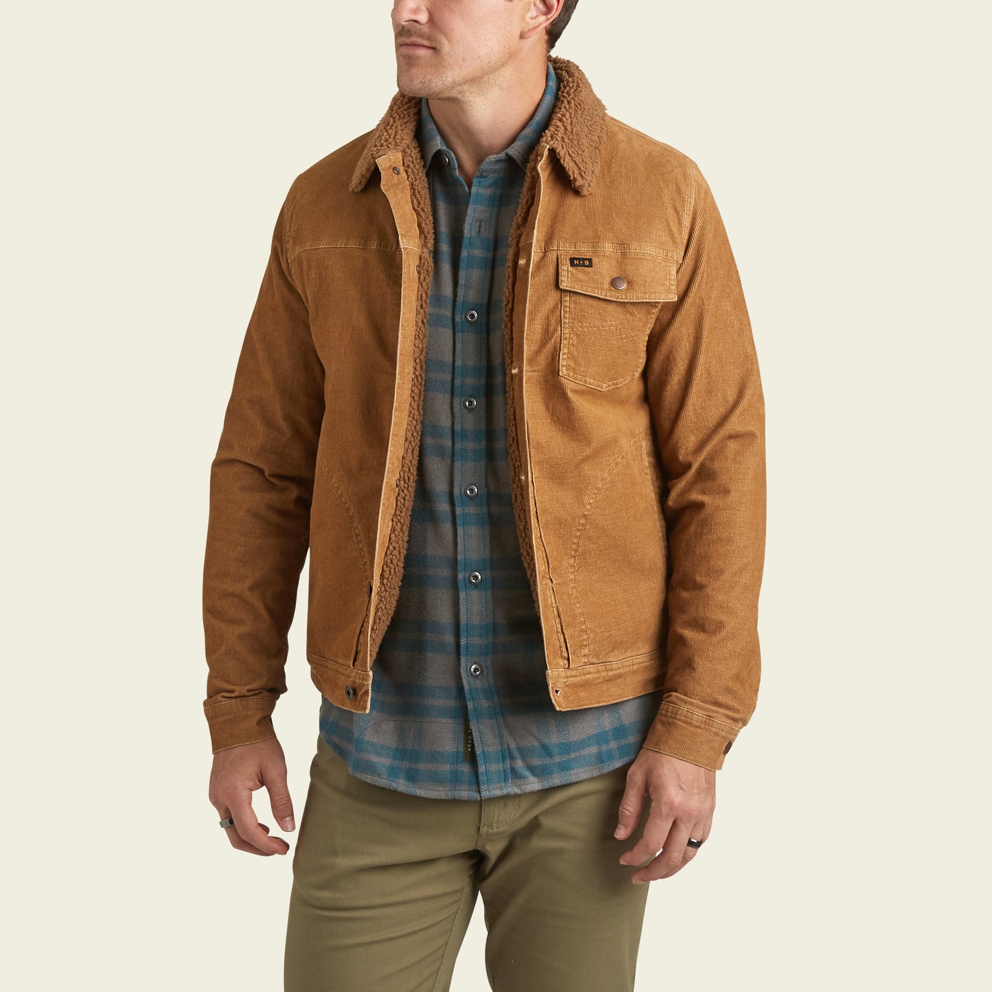 A man wearing a brown Fuzzy Depot Jacket with a sherpa lining over a blue plaid shirt and khaki pants stands facing slightly to the left, with his left hand in his pocket.