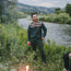 A man in a baja hoodie stands by a riverbank, holding a can and smiling. Lush green bushes and trees surround him, with mountains in the background and a small campfire burning in the foreground.