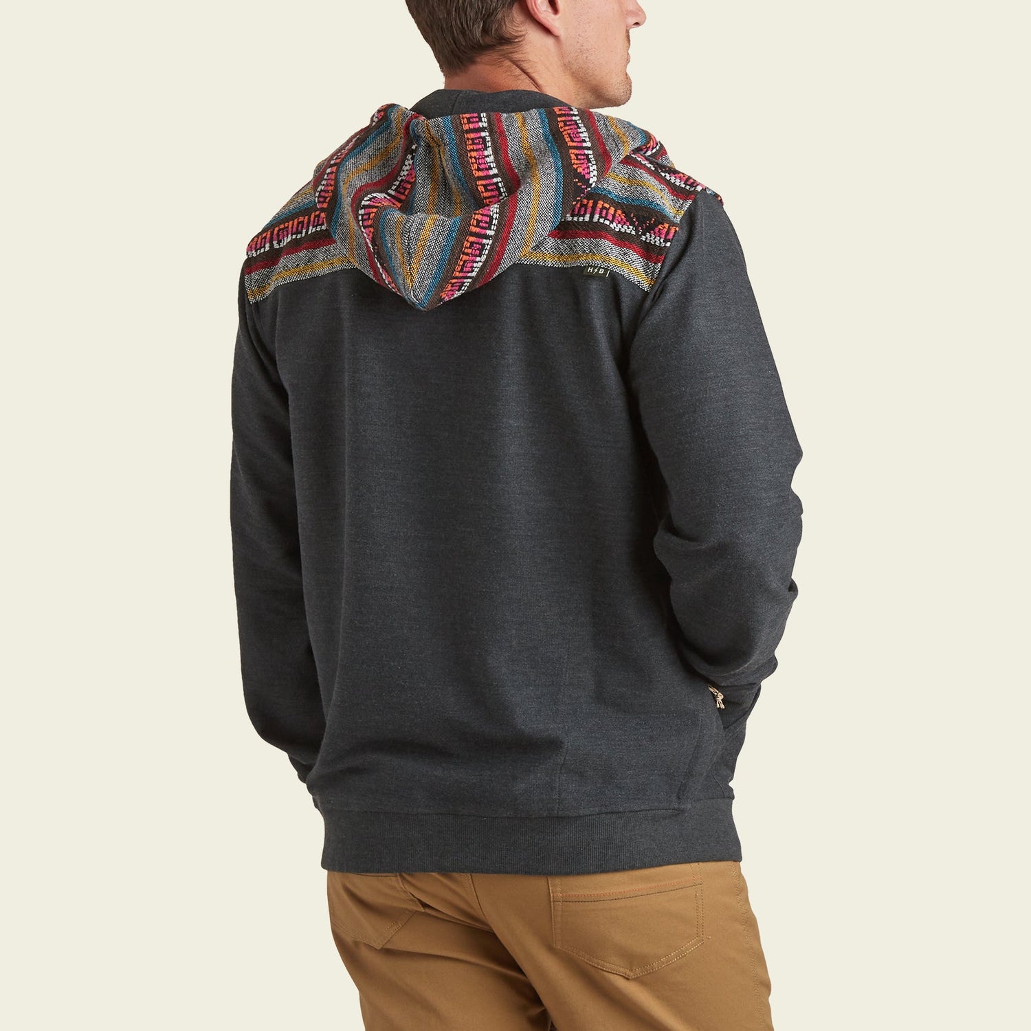 A man wearing a woven jacquard hoodie with colorful, patterned fabric on the hood and shoulders, and tan pants, is standing with his back to the camera.