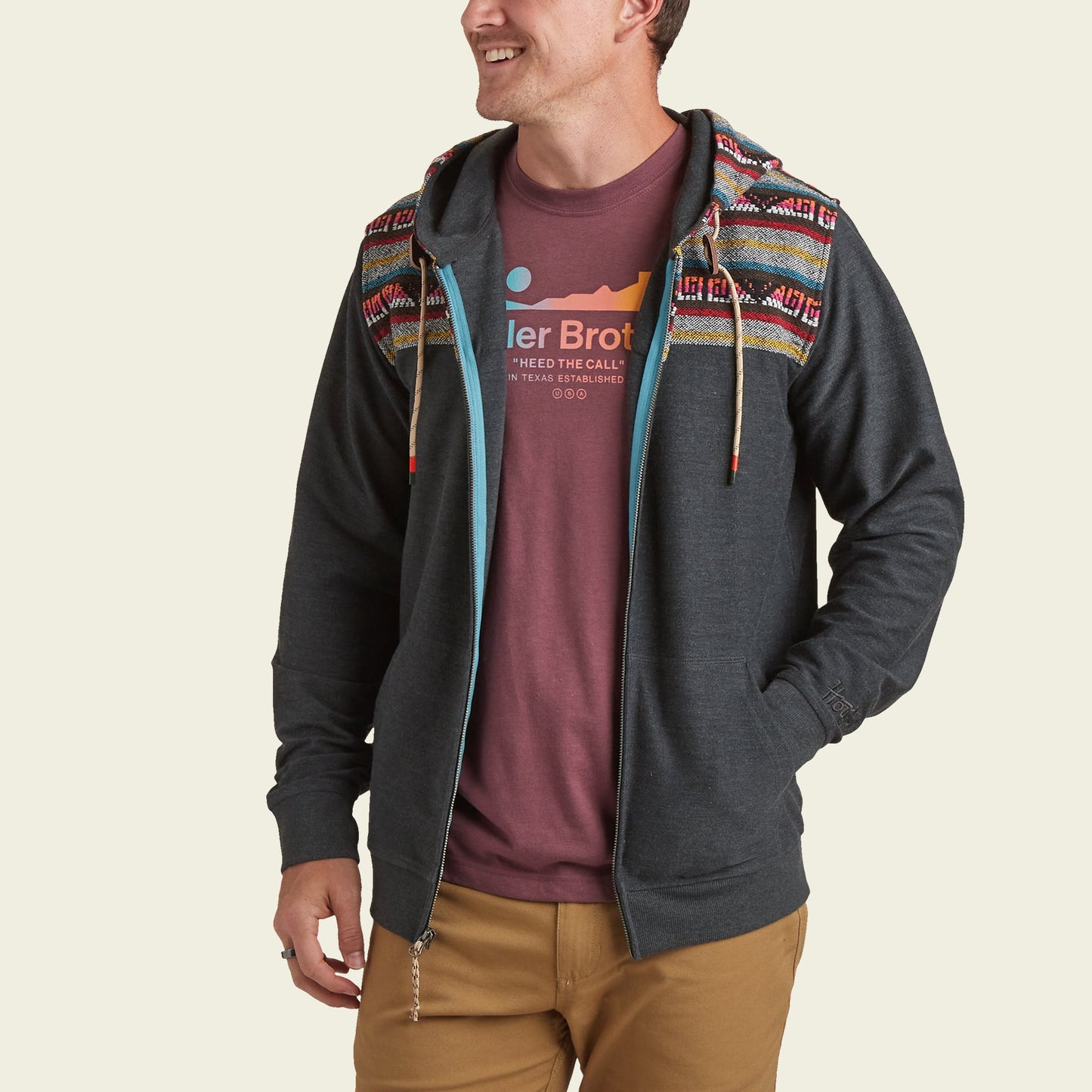 A man smiles while wearing a cotton blend hoodie with colorful patterned shoulders, a maroon graphic t-shirt, and tan pants. His left hand is in his pocket and the hoodie features a light blue lining.