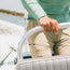 A person wearing a light green long-sleeve shirt and beige tech pants grips a boat steering wheel, with water and a blurred shoreline visible in the background.