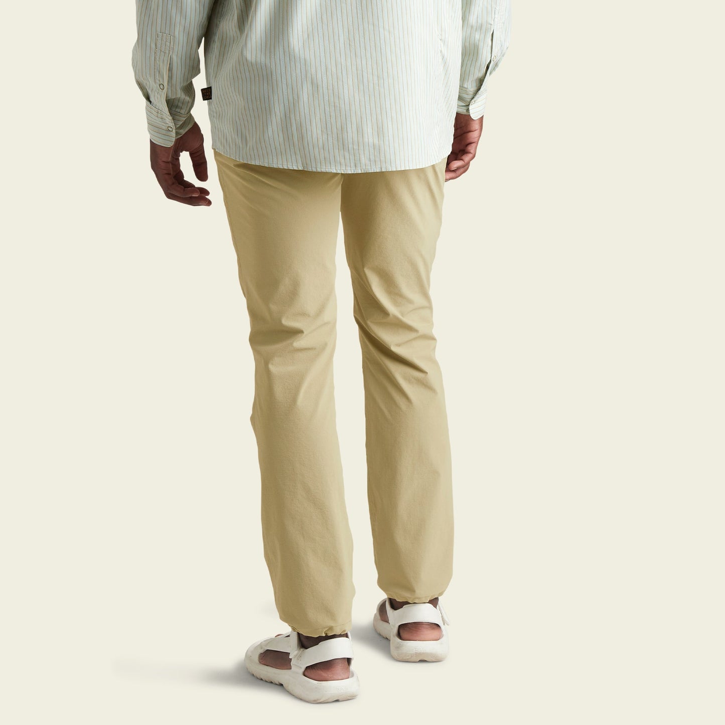 A person wearing light beige tech pants, a light striped long-sleeve shirt, and white sneakers, shown from the back, standing against a plain light background.