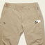Beige tech pants with multiple pockets, including a zippered pocket and a large side pocket holding a white smartphone. These stretch nylon pants feature a button closure and belt loops for comfort and functionality.