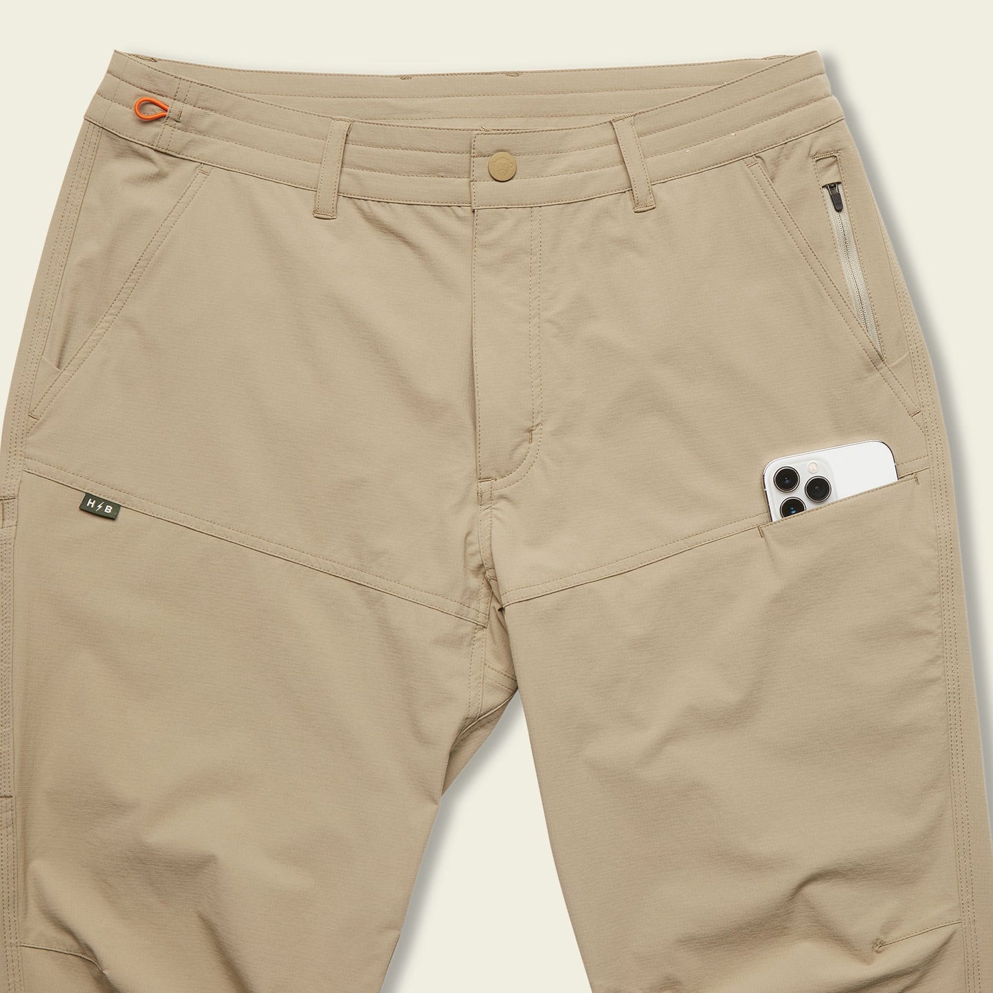 Beige tech pants with multiple pockets, including a zippered pocket and a large side pocket holding a white smartphone. These stretch nylon pants feature a button closure and belt loops for comfort and functionality.