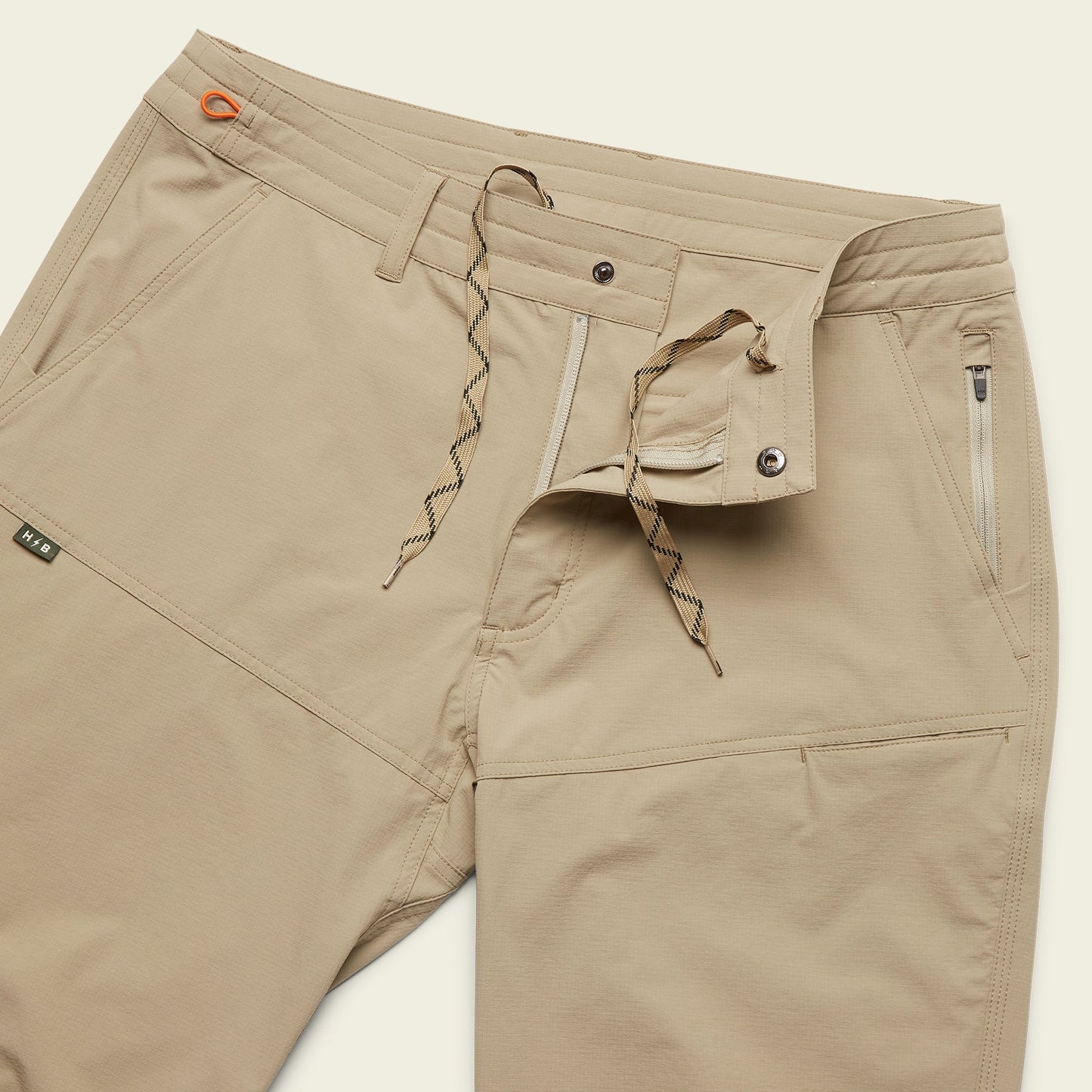 Beige stretch nylon Tech pants with a partially open waistband showing a button, zipper, and drawstring closure. The pants feature front zippered pockets and visible stitching details.