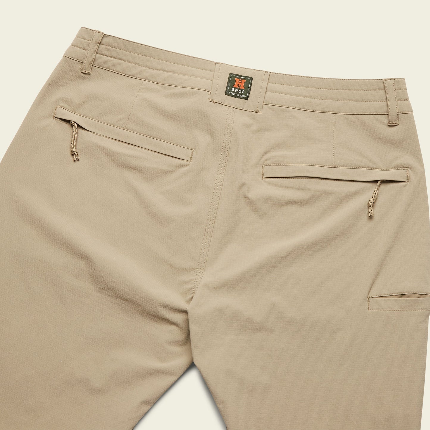 Khaki Tech pants shown from the back, featuring two zippered back pockets, belt loops, and a small stitched logo patch above the right pocket.