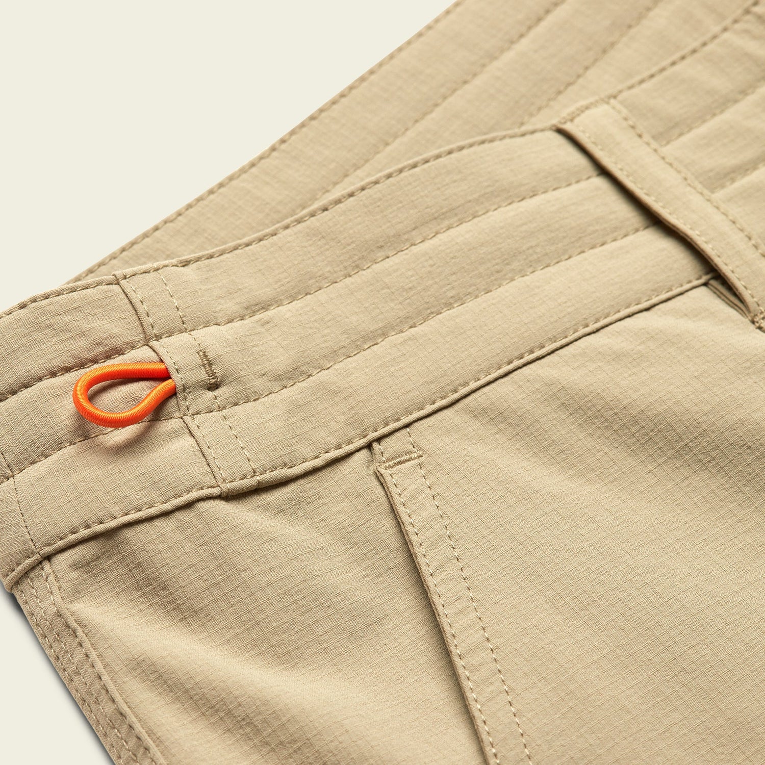 Close-up of tan Tech pants with visible stitching, belt loops, side pocket, and an orange fabric loop detail near the waistband.