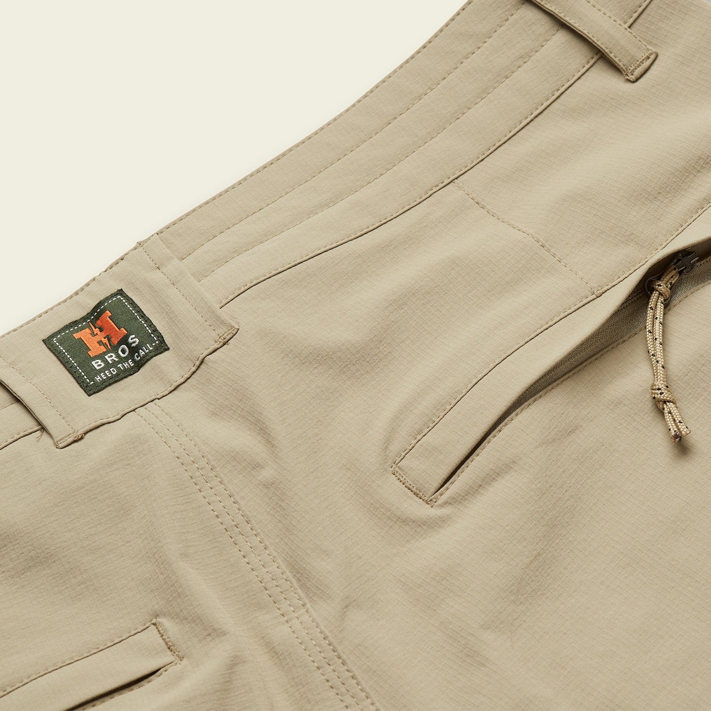 Close-up of beige stretch nylon pants showing a front pocket, belt loops, a drawstring tie, and a green tag with H BROS and PRESTO CHINO printed on it.