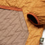 Close-up of a quilted, orange and brown lightweight travel jacket, showing the sleeve, diagonal stitching, and a small green fabric tag with a bear logo on the lower edge.