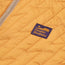 Close-up of an orange quilted pullover jacket with a zigzag stitch pattern, PrimaLoft insulation, a tan zipper, and a rectangular Howler Brothers logo patch in purple and gold.