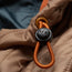 Close-up of an orange elastic drawstring with a black H//B toggle on tan fabric, possibly part of a lightweight travel jacket, with visible stitching and a dark background.