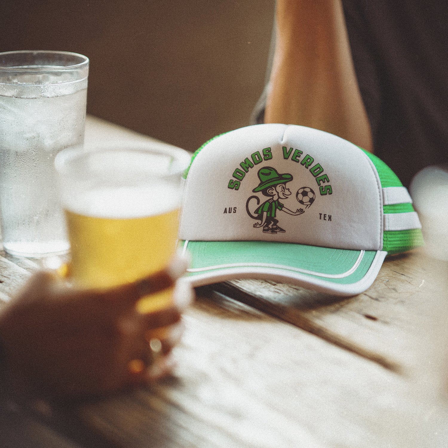 A vintage-inspired hat from the Verde Collection, this green and white trucker displays a cartoon character and SIMOS VERDES AUS TEX, resting beside a glass of water and an Austin FC fan holding a beer.