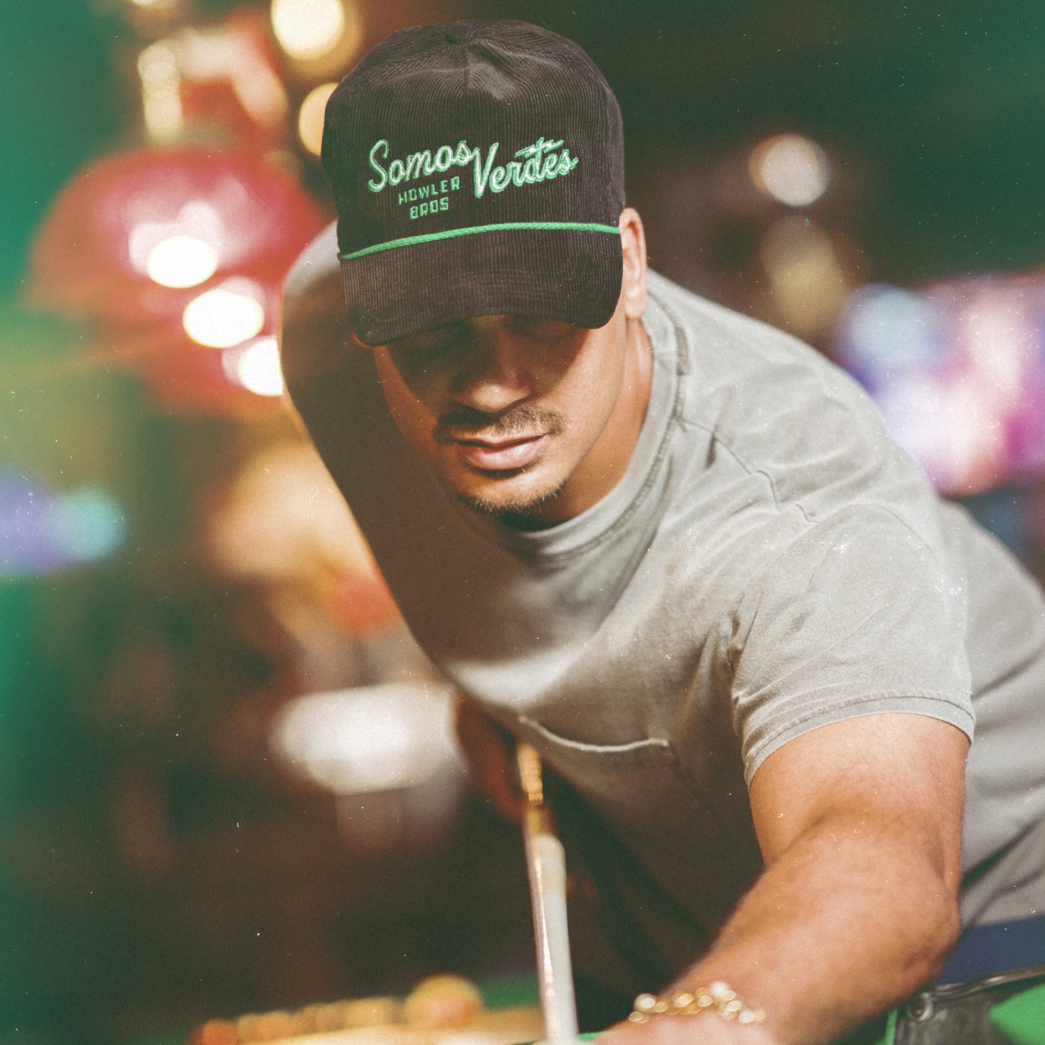 A man wearing a black Austin FC Unstructured Snapback that reads Somos Verdes and a grey t-shirt leans forward, aiming a pool cue in a dimly lit, colorful pool hall.